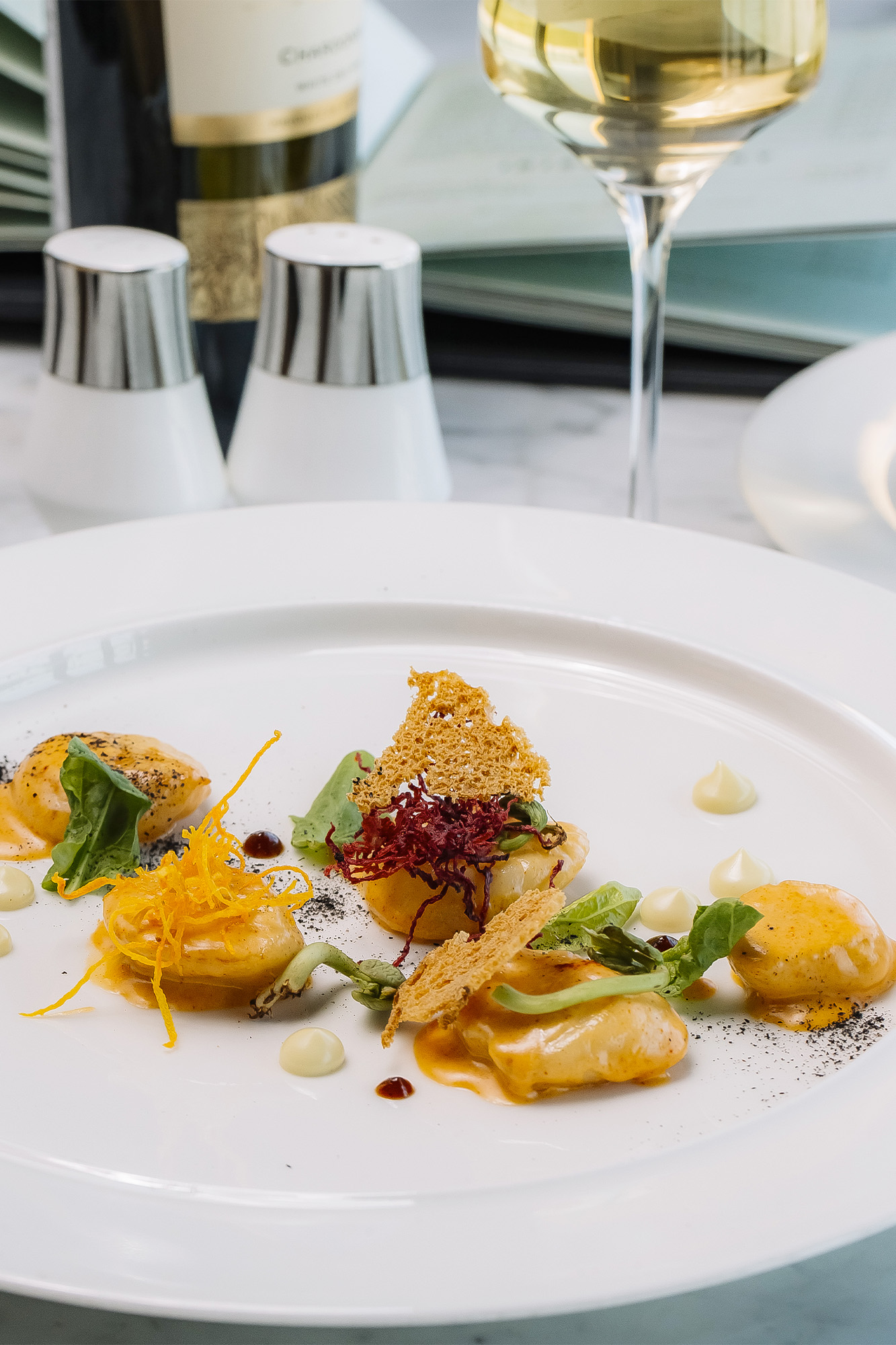 Piatto di ravioli decorato con salsa, erbe e croccante, servito con vino bianco.